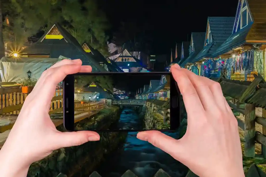 Manos sosteniendo un teléfono móvil mientras capturan una fotografía nocturna de un canal fluvial entre casas iluminadas